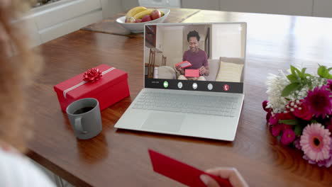 Mujer-Caucásica-Sosteniendo-Un-Sobre-Rojo-Usando-Una-Computadora-Portátil-Con-Un-Hombre-Afroamericano-Con-Regalo-En-La-Pantalla