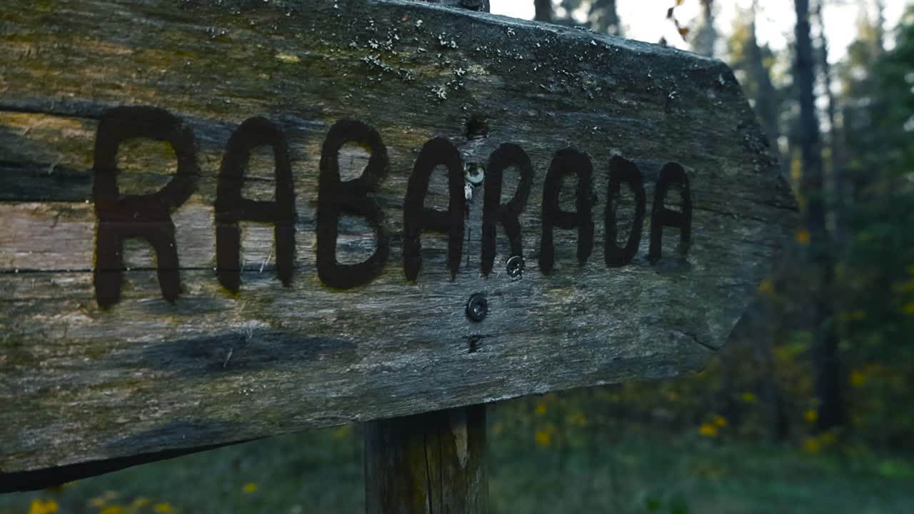 Close cinematic glide toward weathered wooden Rabarada trail sign pointing to bog path surrounded by autumn forest with soft blurred foliage