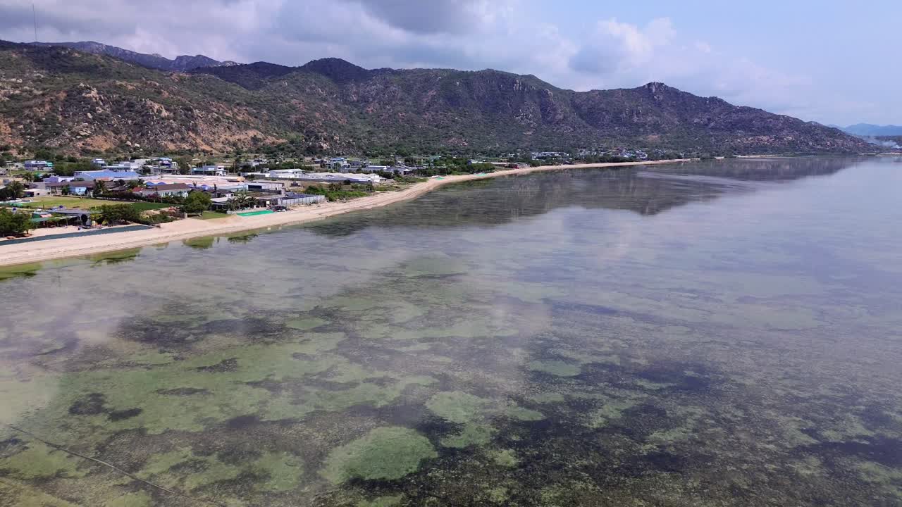 drone flies forward with an upward pan, revealing the beach on the left and ahead, distant mountains, and a clear horizon in Vietnam