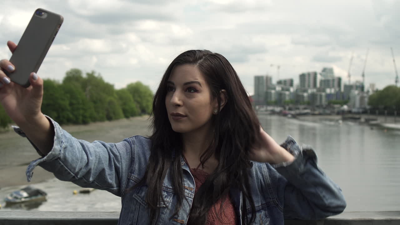 hermosa latina divirtiéndose mientras se toma una selfie con su teléfono, posa, guiña un ojo y se lo pasa genial mientras está de pie en un puente con vista a londres