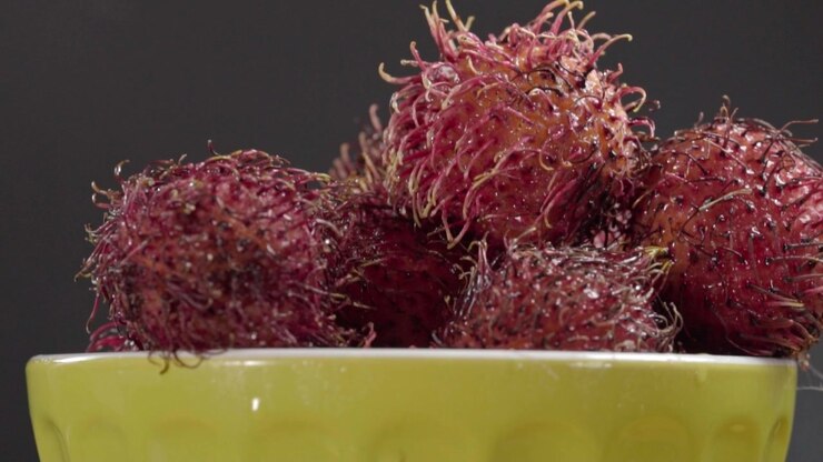 Frutti di rambutan freschi con buccia rossa pelosa disposti in una ciotola, che mostrano le loro caratteristiche spine curve e superfici texturizzate su uno sfondo scuro