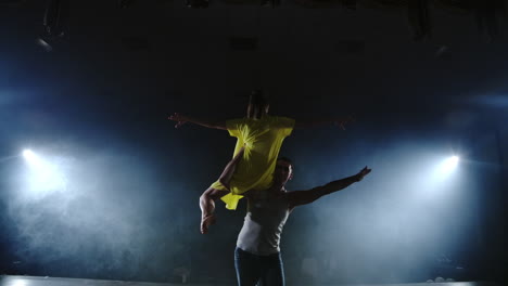 Zoom-camera-a-female-dancer-jumps-onto-her-partner-shoulder.-Acrobatic-dance-musical-on-stage-in-smoke.-Contemporary