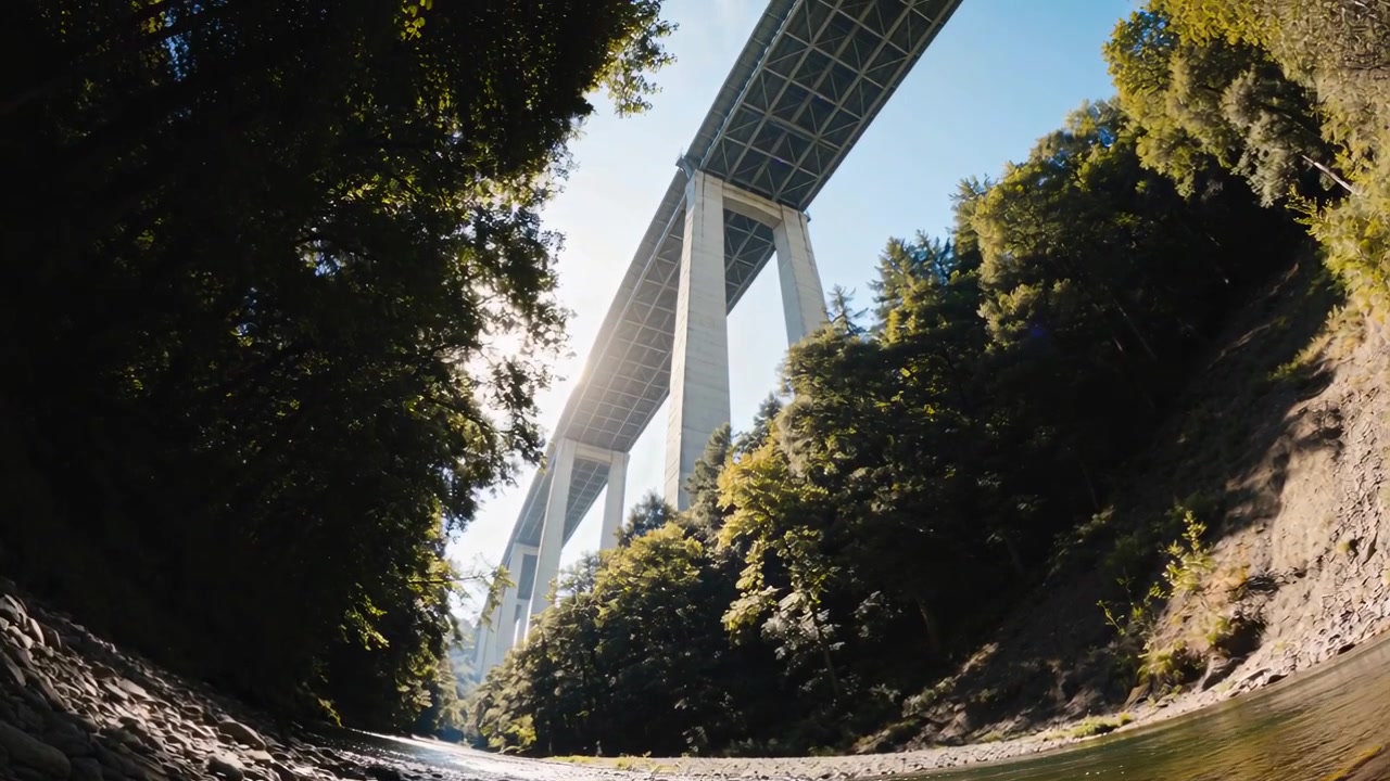 Câmera inclinando-se para baixo após o operador empurrar o tripé, revelando um viaduto de concreto sobre um riacho iluminado pelo sol.