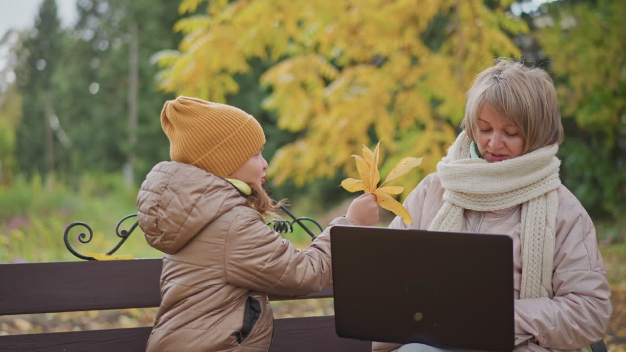 woman sits on bench working laptop outdoors in autumn park while daughter approaches holding yellow leaves to distract mother who gently shifts hand aside before child retreats