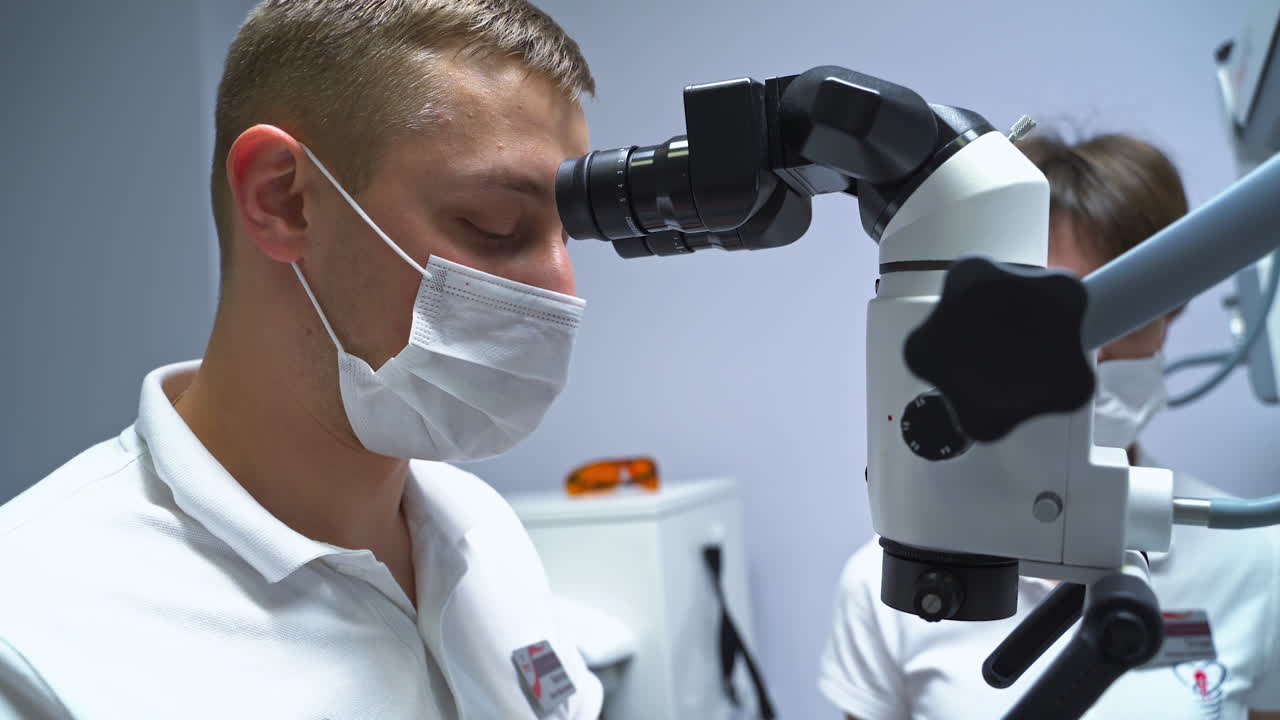 Dental care specialist working with a patient looking into equipment binoculars. Handsome young male doctor using tools and technology. Nurse at backdrop.
