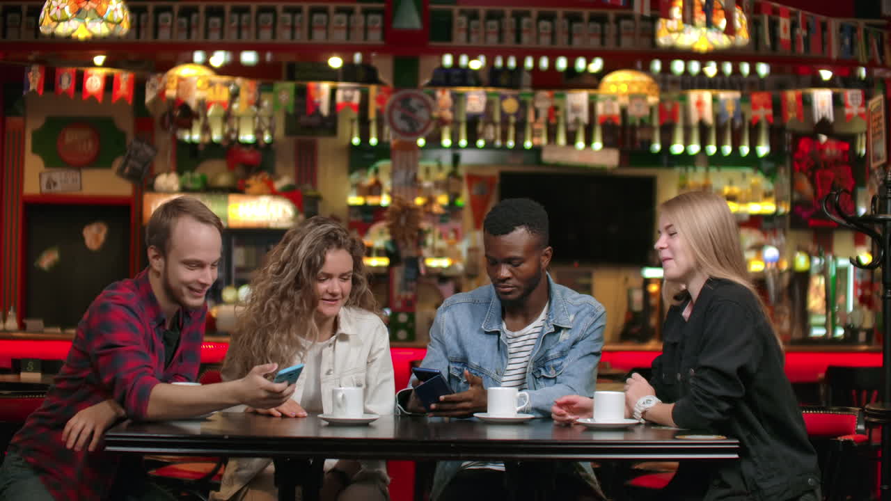 Four friends look at photos on the phone and laugh while sitting in a coffee shop and discussing what they saw on the screen
