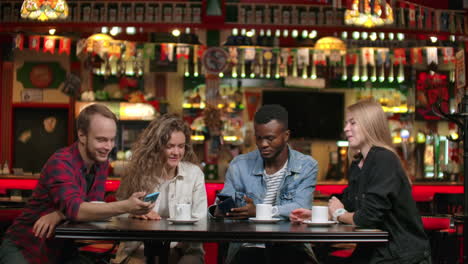 Vier-Freunde-Schauen-Sich-Fotos-Auf-Dem-Telefon-An-Und-Lachen,-Während-Sie-In-Einem-Café-Sitzen-Und-Darüber-Diskutieren,-Was-Sie-Auf-Dem-Bildschirm-Sehen