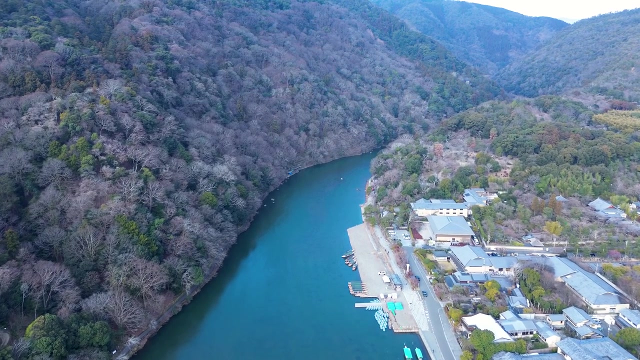 日本の京都、嵐山公園亀山地区。山間の谷沿いに広がる鬱蒼とした森林や低木、町や村の家々に囲まれた桂川の水路をドローンで空撮。自然と観光の風景。