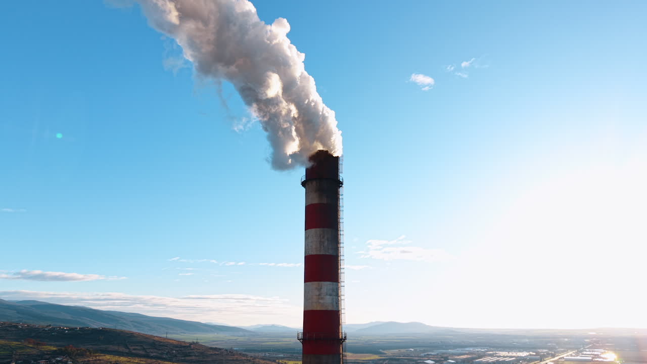 Il fumo sale dal camino di una fabbrica in un cielo azzurro sopra le colline. Un camino di una fabbrica rilascia fumo nel cielo