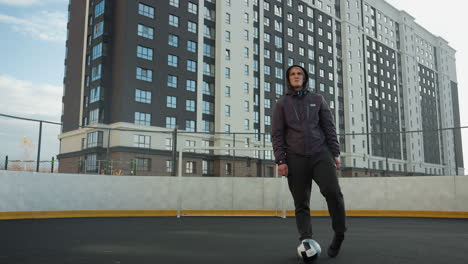 sportsman performing ankle twists with left and right legs, balancing ball in front on urban sports arena with goalpost and tall urban building in background