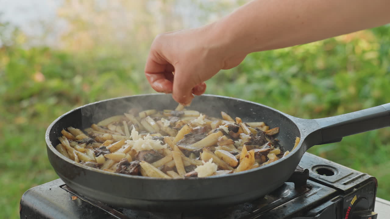 Padella da campeggio cosparsa di formaggio grattugiato su patate e funghi, mano che aggiunge l'ultima guarnizione sopra la padella fumante su fornello portatile vicino al lago, calda luce dorata, una confortante esaltazione del sapore dal fresco.