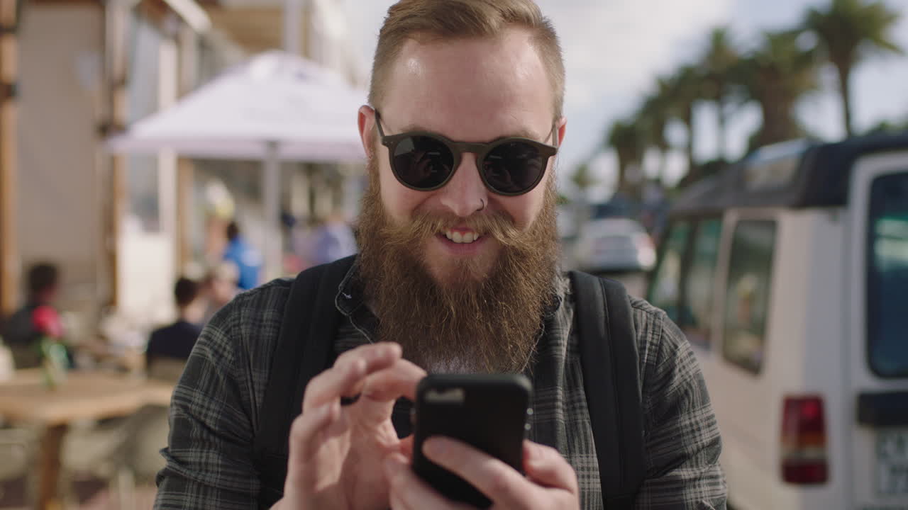 portrait of bearded hipster man wearing sunglasses using phone texting on busy beachfront