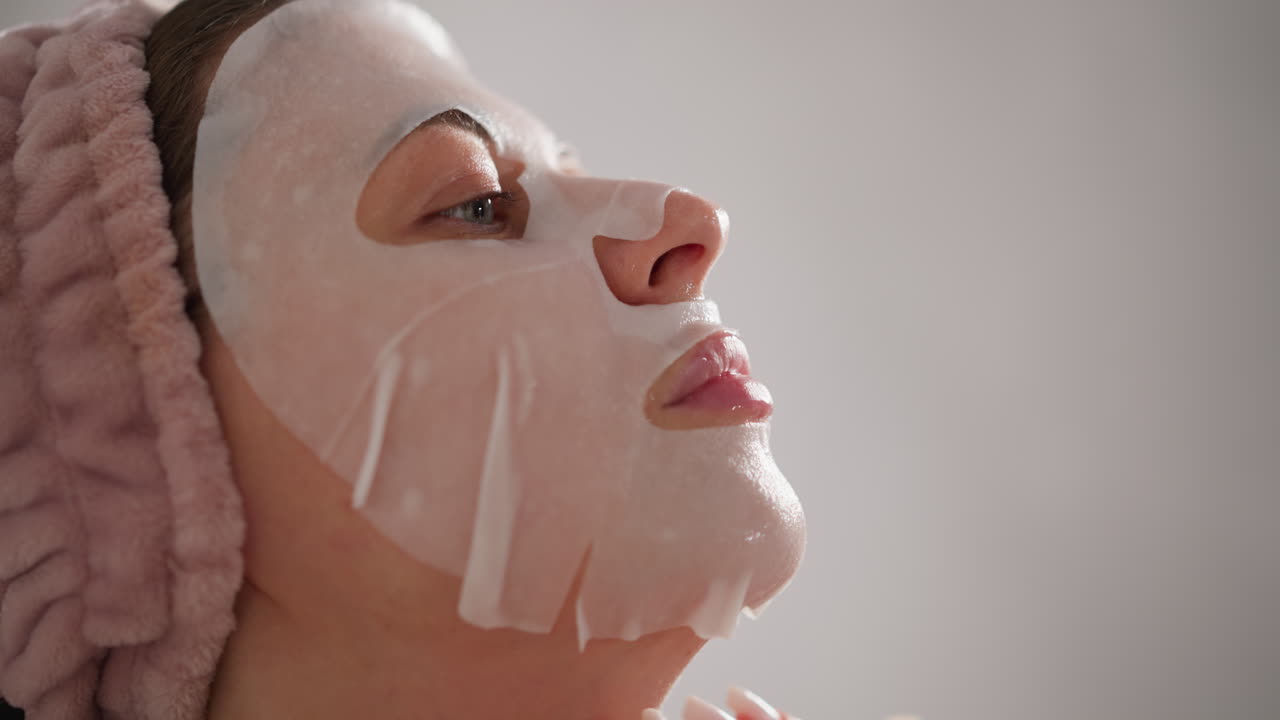 여성은 화장실에서 얼굴에 화장품 천 마스크를 매끄럽게합니다. 여성은 힘든 일 후에 얼굴 마스크를 만드는 즐거운 화장품 절차에 참여합니다.