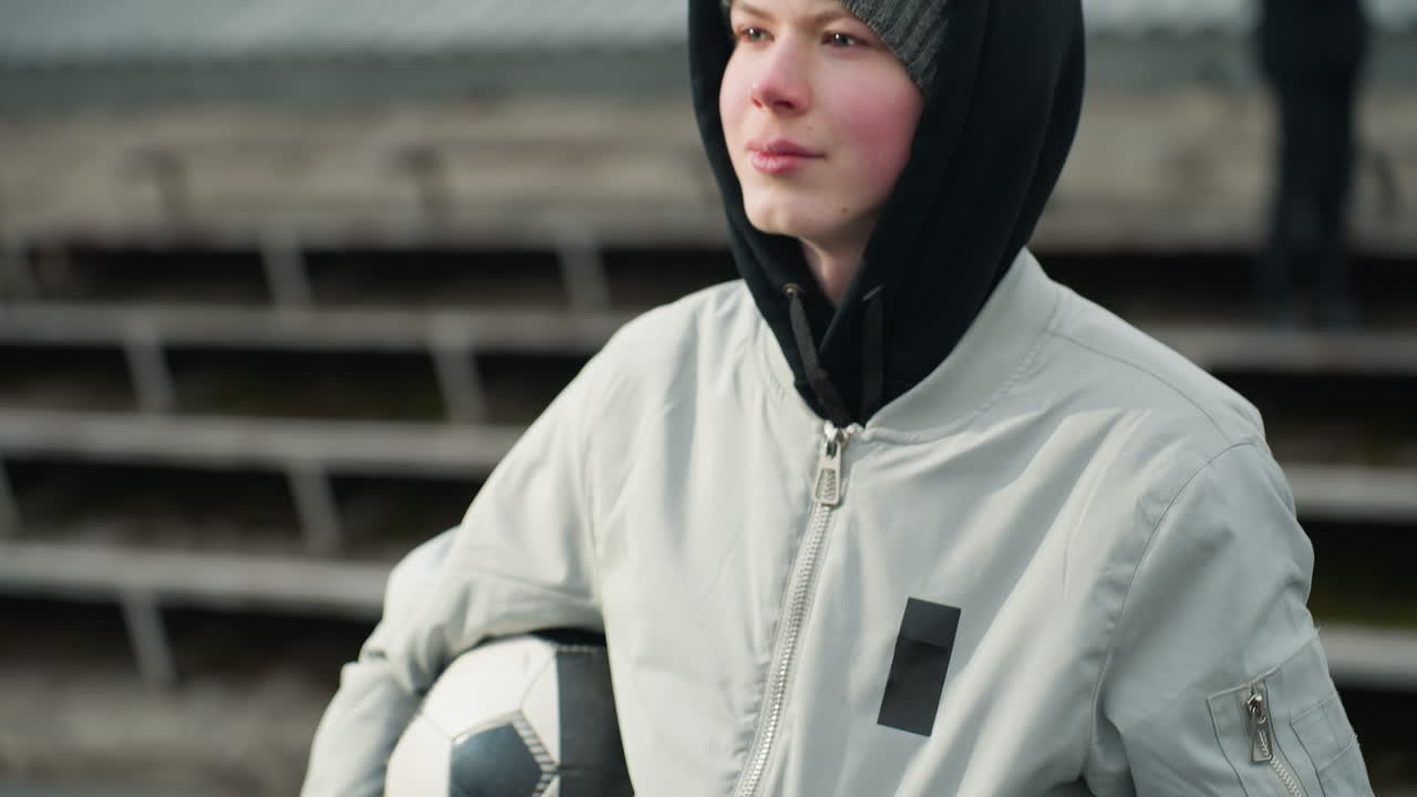 Close-up of a young boy wearing a gray sweater, holding a soccer ball under his arm while looking focused