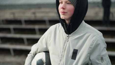 close-up of a young boy wearing a gray sweater, holding a soccer ball under his arm while looking focused