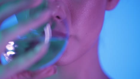 vista de cerca de una mujer joven con labios hermosos llenos bebiendo un vaso de agua para saciar su sed contra un fondo azul con contraste púrpura en su cara en cámara lenta