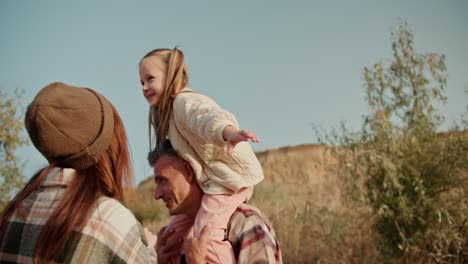 a little blonde girl has fun when she sits on the shoulders of her dad, the brunette man with a little gray hair in a checkered shirt, and talks to her mom, the brunette girl in a green checkered shirt, during her picnic