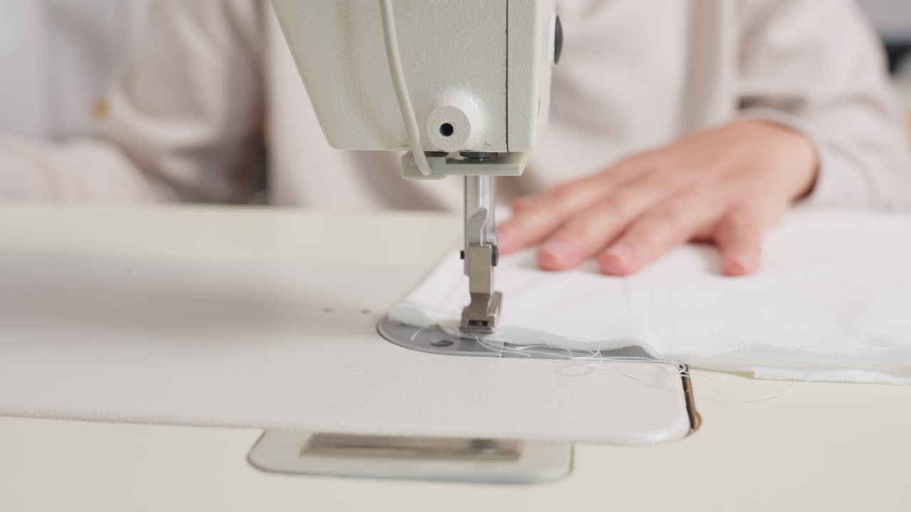 Electric machine needle sewing through white cotton fabric as stylist guides material forward, ensuring alignment, consistent tension, demonstrating skilled technique, focusing on finishing precision