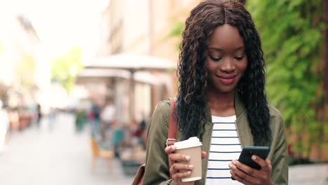 african american woman holding coffee to go and using smartphone while walking down the street