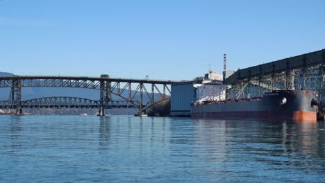 Cargo-ship-docked-at-industrial-port-in-Vancouver,-sunny-day-with-clear-blue-sky,-bridge-in-the-background,-calm-waters