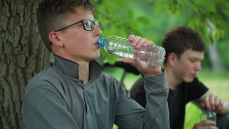 two young siblings resting under a tree with one of them drinking water while the other bows his head, they are taking a break from a bike ride, with bicycles parked nearby surrounded by greenery