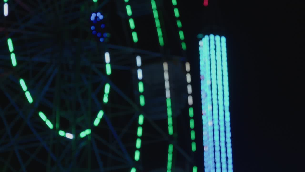A dreamy, abstract shot of a carnival at night. The green and blue lights of a Ferris wheel and a tower ride are blurred into a beautiful bokeh background