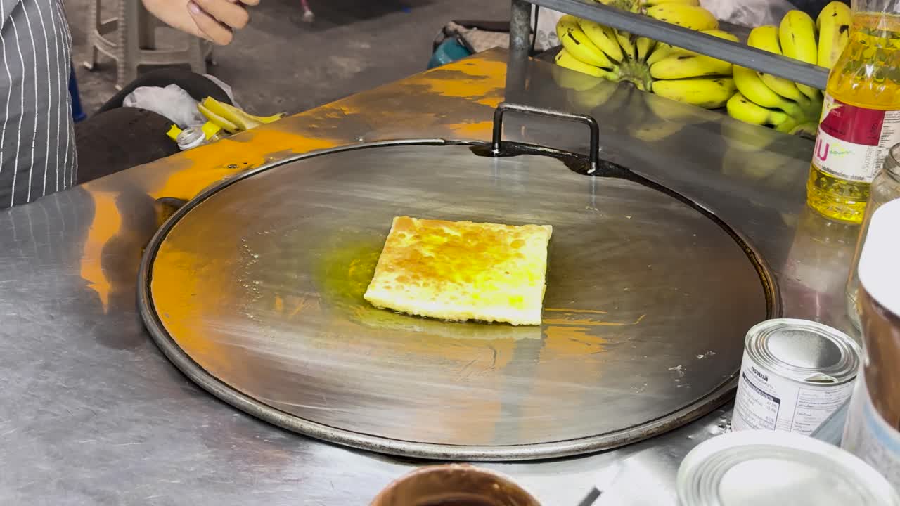 un vendedor callejero prepara hábilmente roti tailandés en una parrilla caliente, mostrando técnicas de cocina tradicionales en un entorno de mercado vibrante