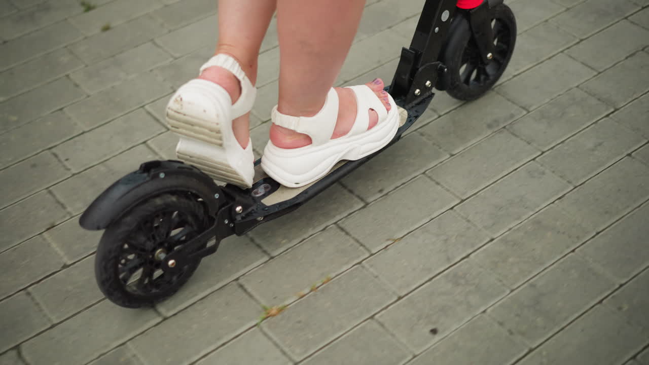색 샌달을 입고 검은색 스쿠터를 타고 있는 여성의 근접 다리 시각, 그녀는 서로 연결된 경로에서 부드럽게 미끄러지고 있습니다.