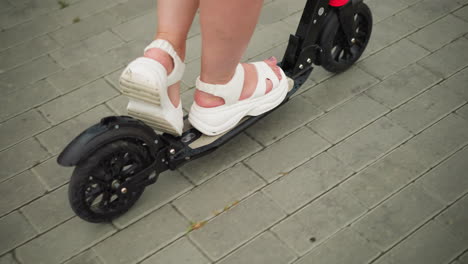 close-up leg view of a woman riding a black scooter while wearing white sandals, she is gliding smoothly on an interlocked pathway