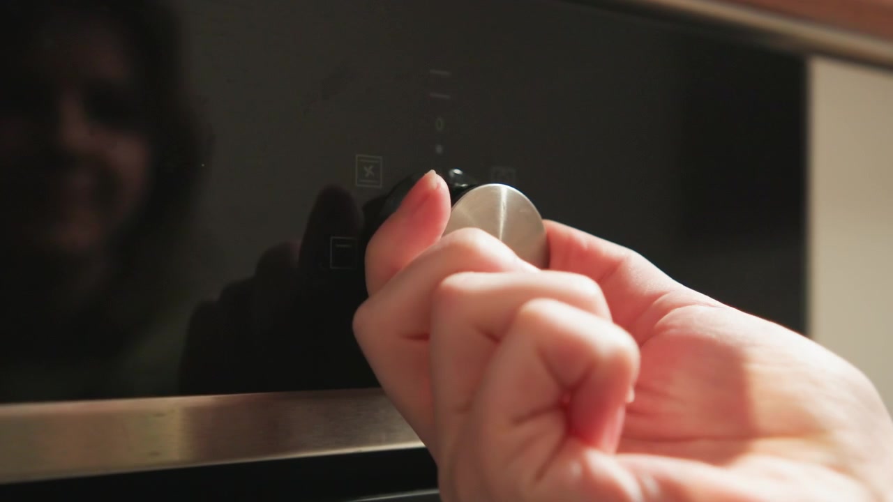 Primo piano delle mani che afferrano e girano una manopola di controllo in acciaio inossidabile sulla parte anteriore lucida e nera di un forno, mostrando il movimento di torsione, il contatto delle dita, la superficie riflettente e una regolazione attiva in cucina.