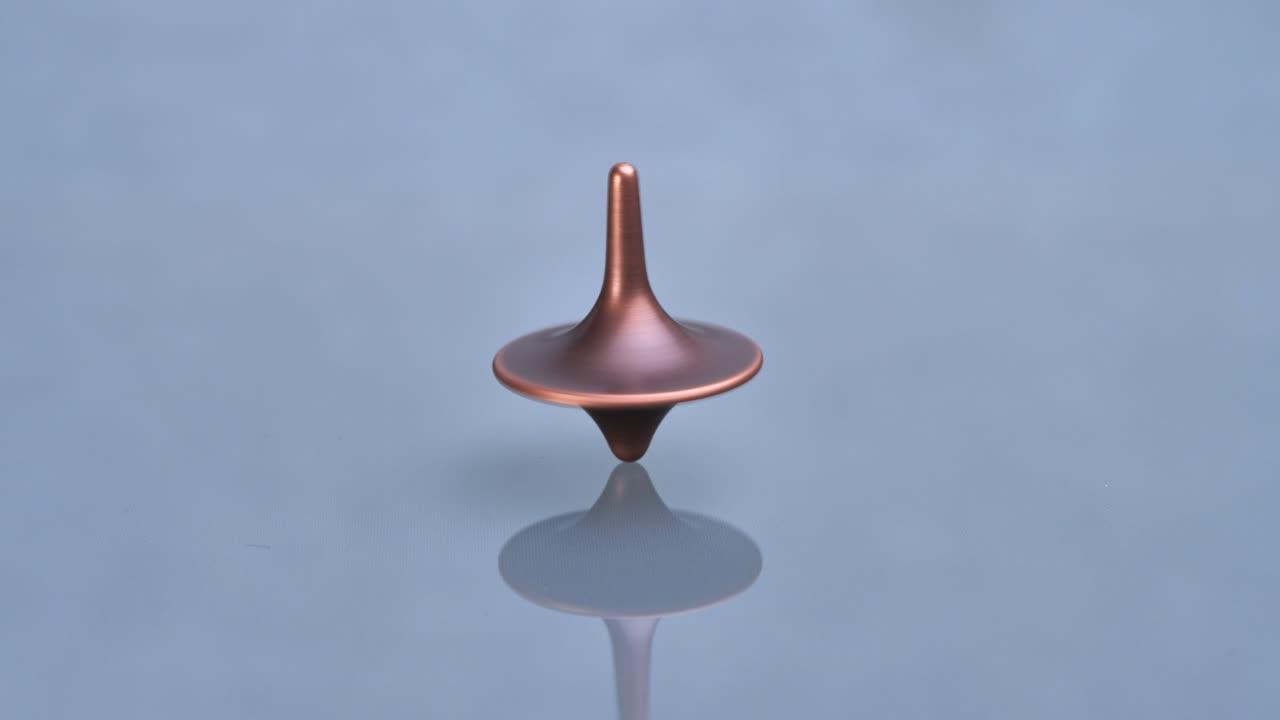 A copper-colored, metallic spinning top (totem) spins on a reflective, gray-blue surface with a soft, mirrored reflection