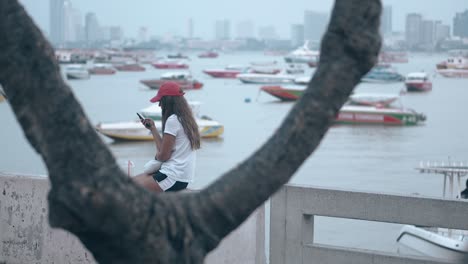 lady-in-red-cap-reads-sms-in-phone-sitting-against-sea-bay