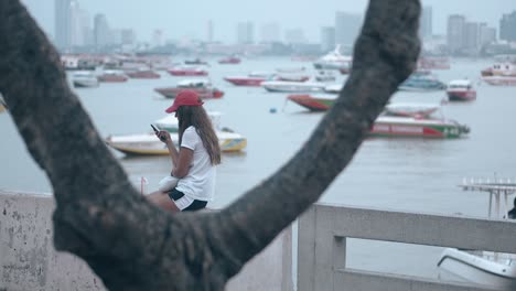 lady in red cap reads sms in phone sitting against sea bay