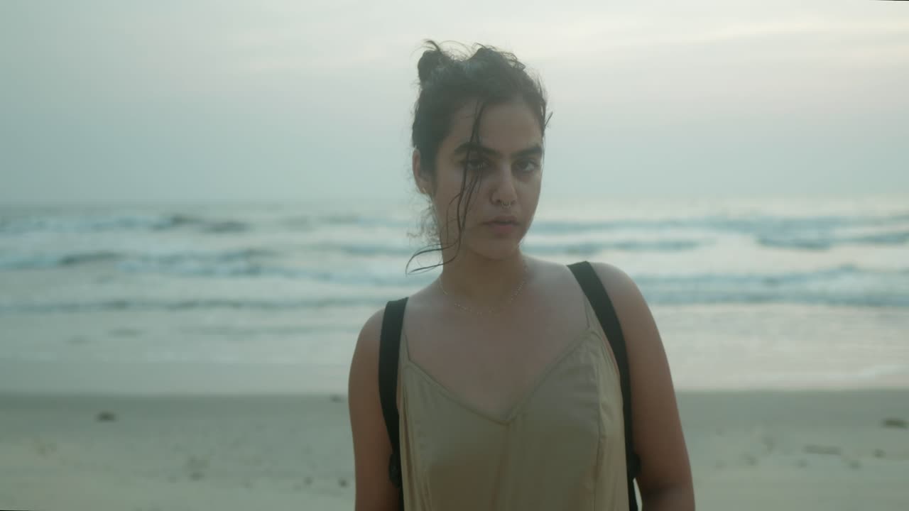 Donna indiana in piedi su una spiaggia al tramonto, che guarda intensamente la telecamera