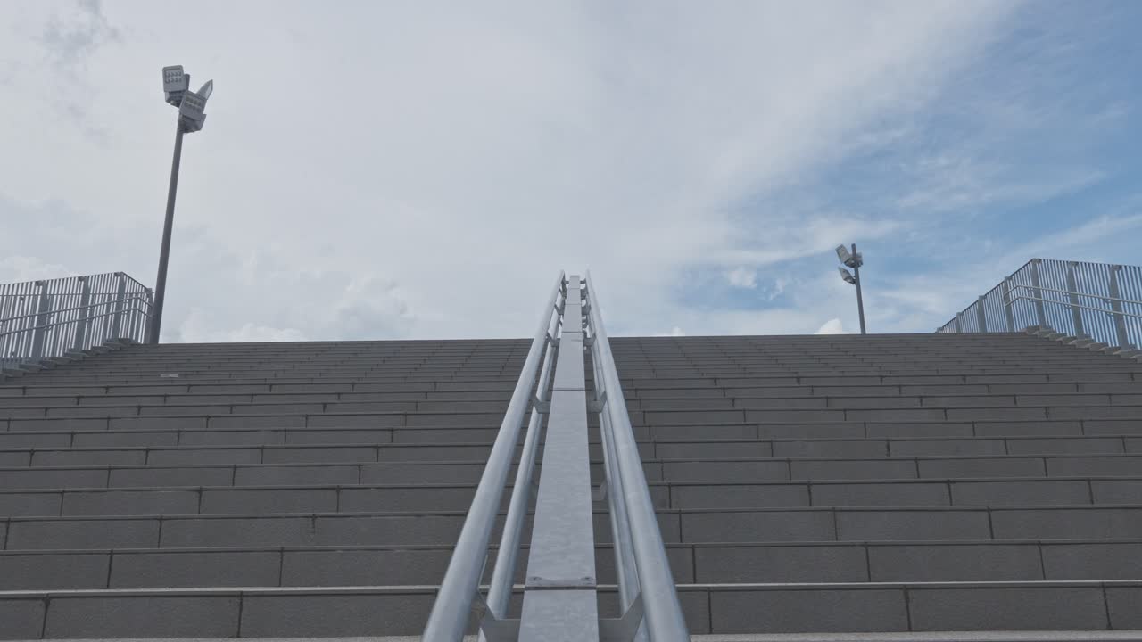 A low-angle shot of a wide concrete staircase with metal handrails leading up to a cloudy sky