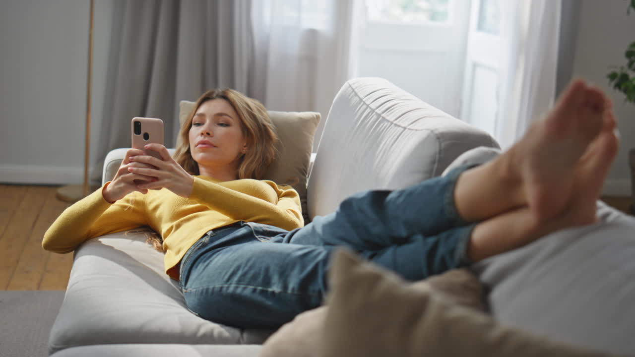 Laying model watching mobile phone screen at comfortable sofa. Chilling woman