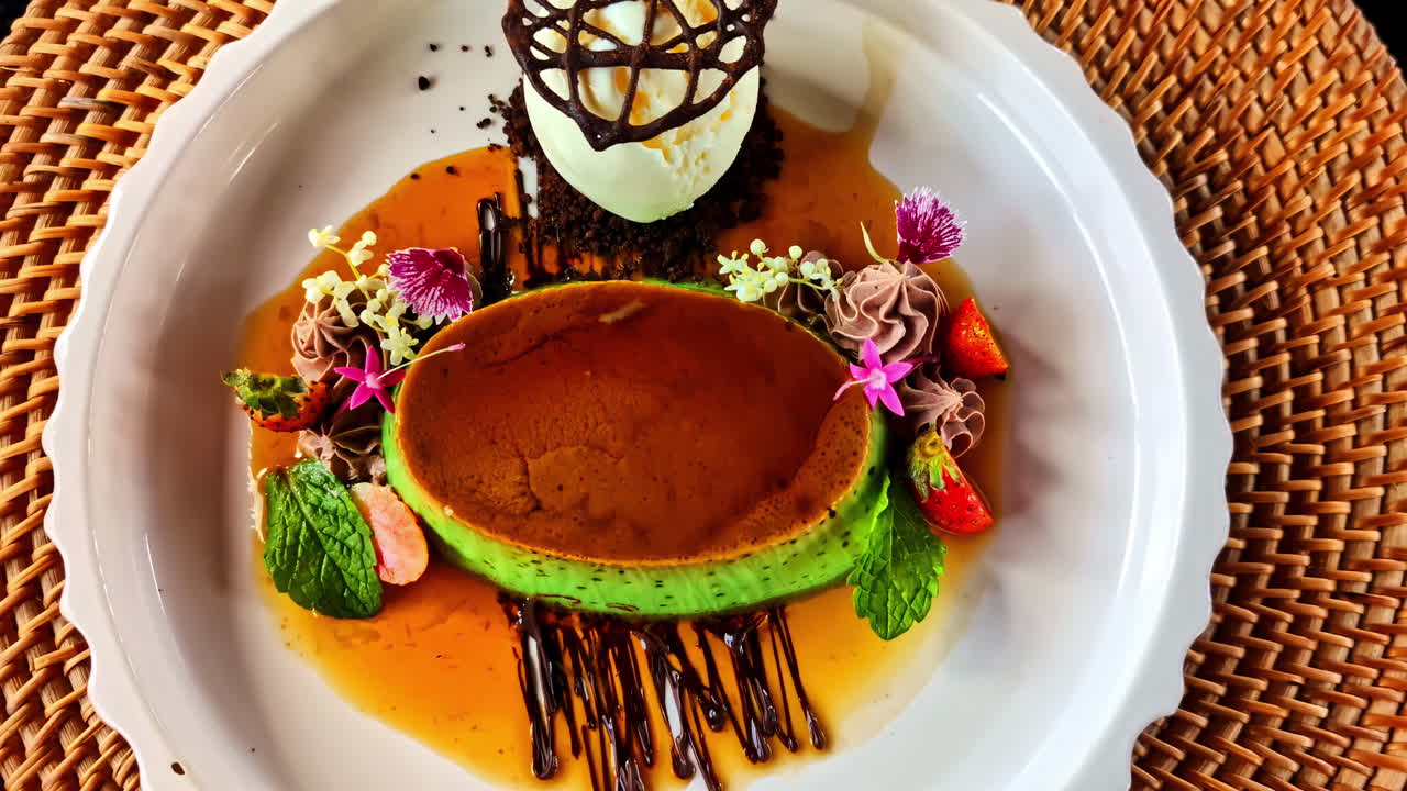 An exquisitely plated gourmet dessert featuring a pandan crème brûlée with caramel, a scoop of vanilla ice cream in a chocolate cage, and decorated with edible flowers at Kuta Selatan Bali