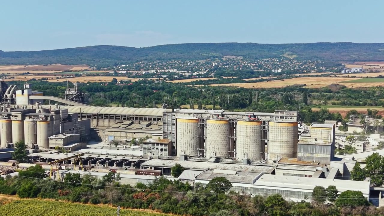 Arbeiter verrichten Aufgaben auf einem großen Industriegelände nahe einem ländlichen Gebiet mit Feldern und Hügeln im Hintergrund