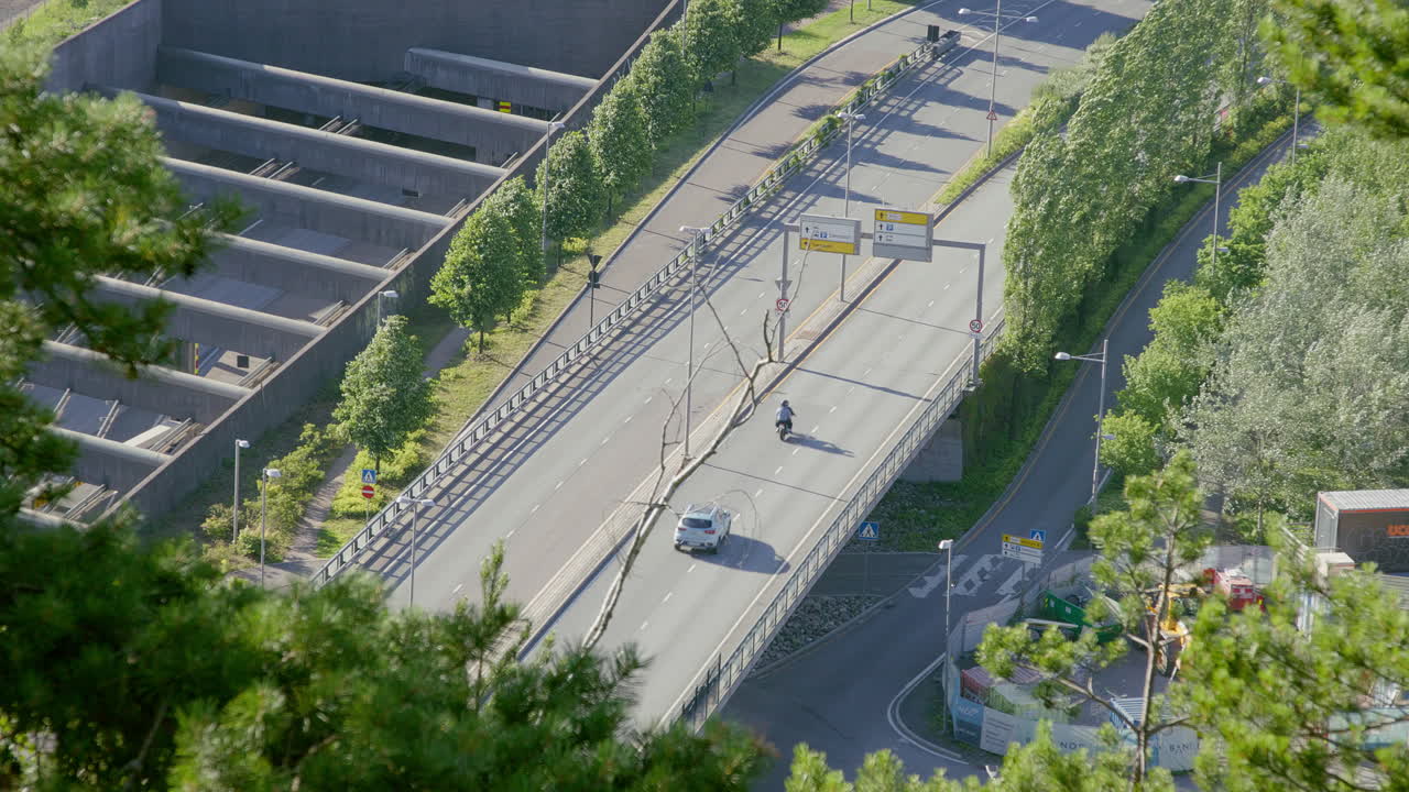 Aerial perspective of a highway with vehicles, bordered by trees and modern urban structures. Filmed on a sunny summer day.