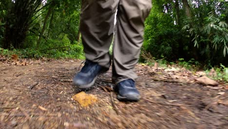 A-man-walking-in-the-jungle,-camera-focused-on-his-legs,-showing-him-stepping-forward-on-the-dirt-road-with-his-shoes-framed