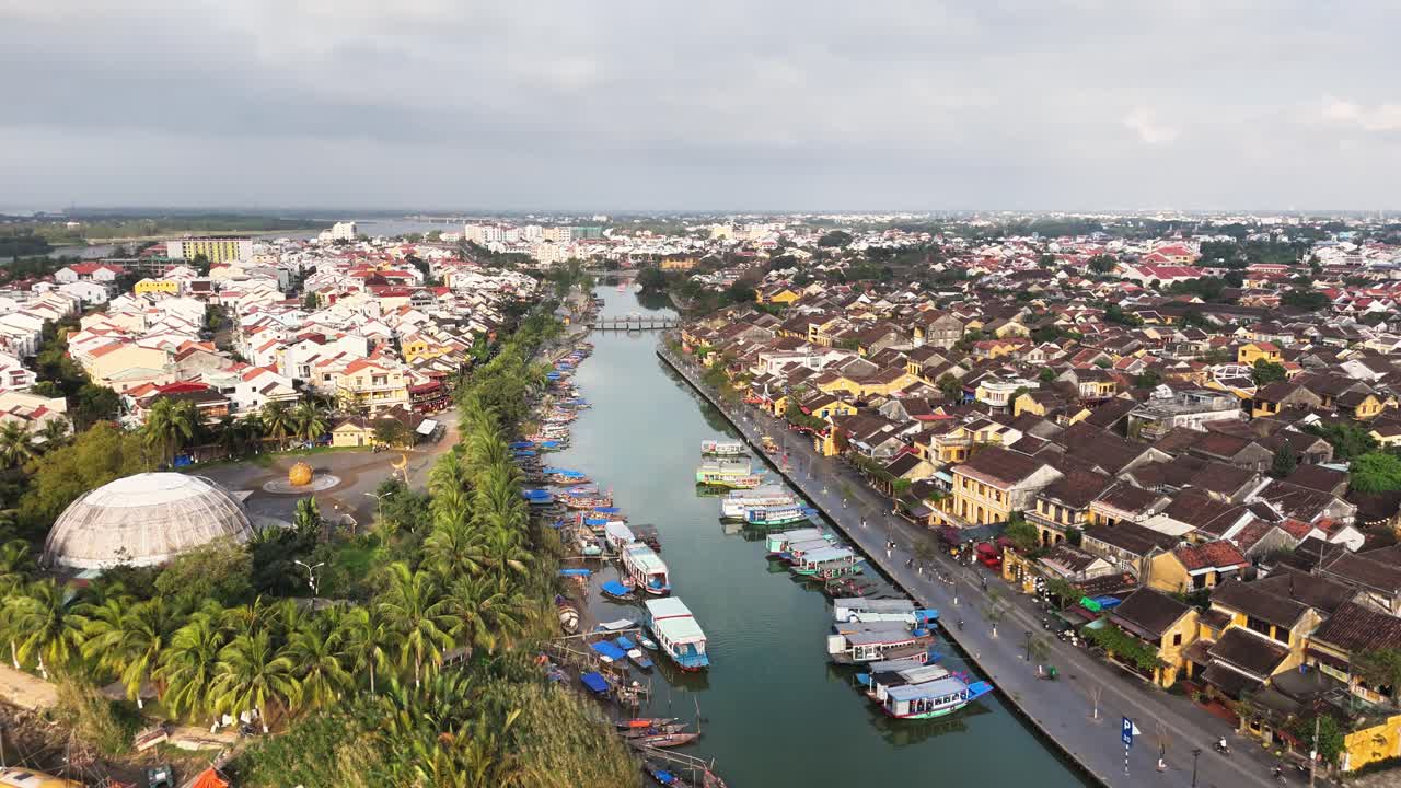 Aerial view of Hoi An, Vietnam, featuring a serene river lined with boats, traditional yellow-tinted buildings, palm trees, and a blend of historic charm with vibrant urban surroundings