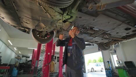 joven mecánico de automóviles en una estación de servicio de reparación inspeccionando la rueda del automóvil y el detalle de la suspensión de un automóvil levantado. vista de abajo.