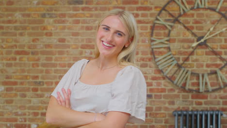 portrait of confident young woman standing by brick wall with clock at home smiling at camera