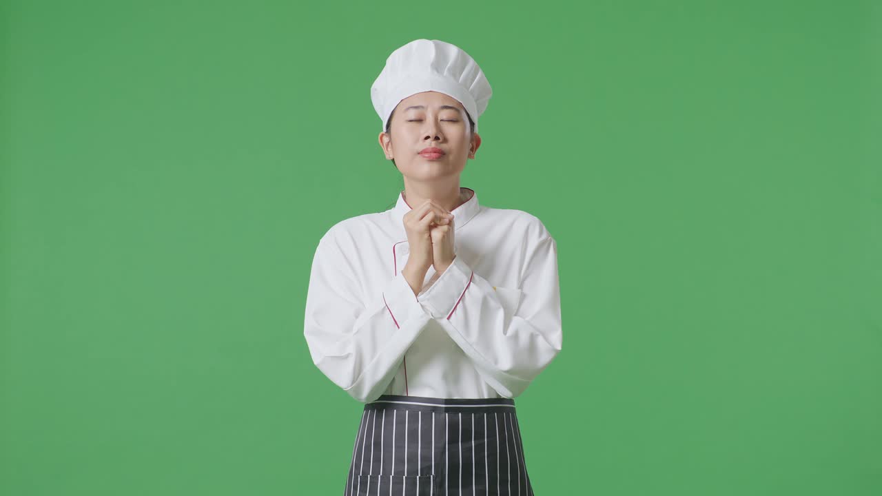녹색 스크린 배경 스튜디오에 서 있는 동안 무언가를 위해 기도하는 아시아 여성 요리사
