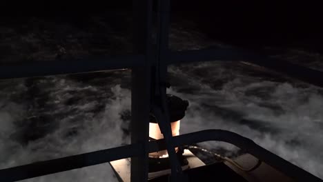 close-up-of-yellow-safety-light-on-ferry-ship-as-it-sails-through-the-dark-night,-and-white,-rough-black-and-white-ocean-waves-pass-by