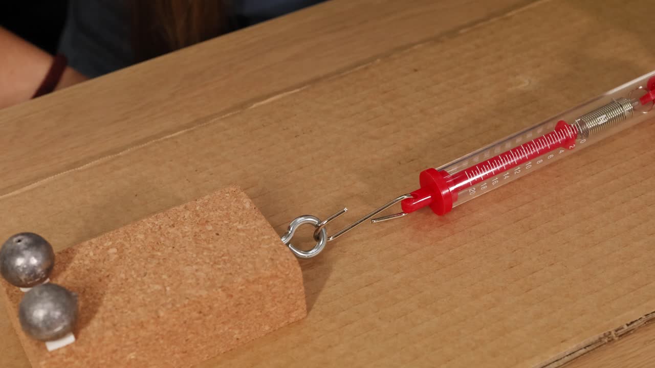 A spring scale measures force as a cork block is pulled across cardboard, illustrating friction and weight dynamics