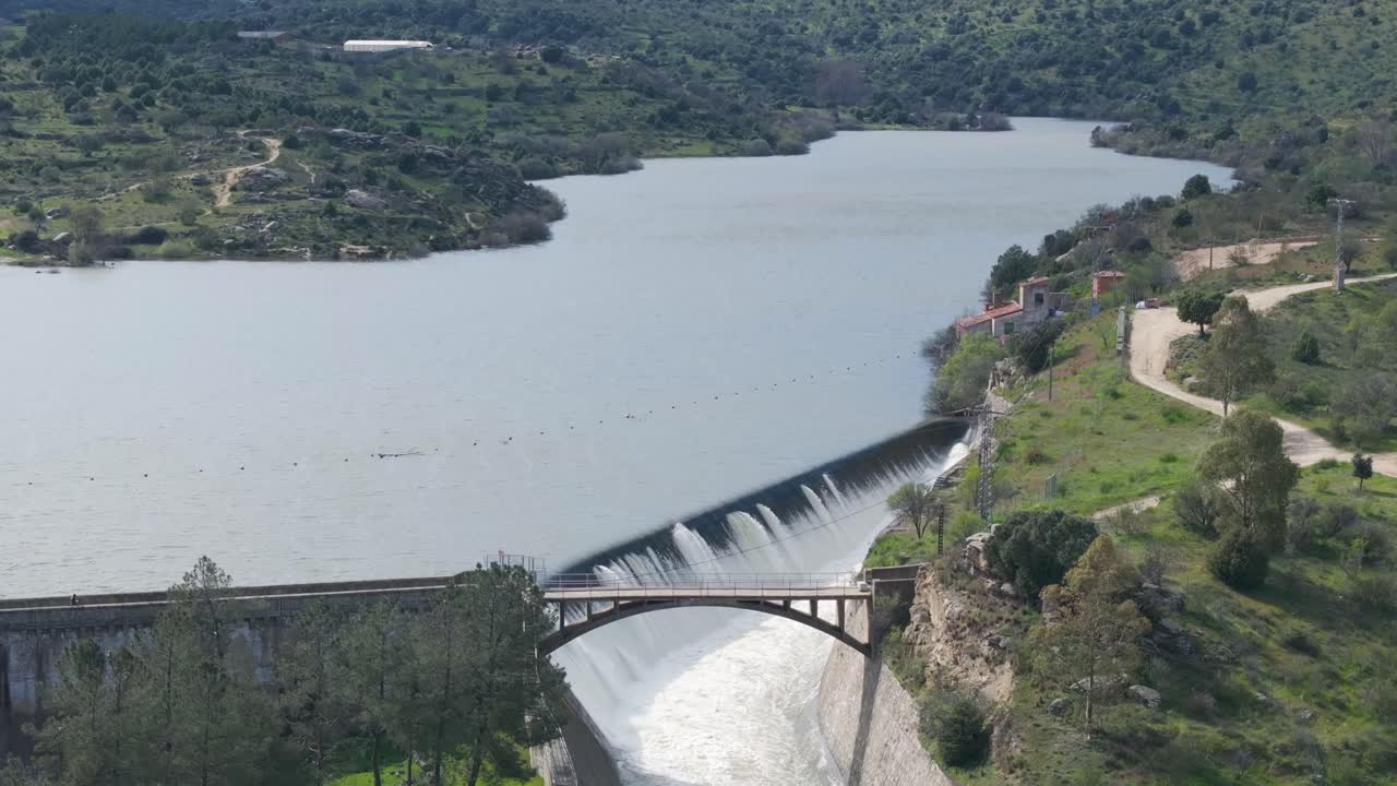 Static aerial shot in front of a dam overflowing through the spillway with incredible force. The rushing water falls beneath a bridge built directly over the powerful outflow channel.