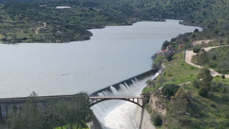 static aerial shot in front of a dam overflowing through the spillway with incredible force. the rushing water falls beneath a bridge built directly over the powerful outflow channel.
