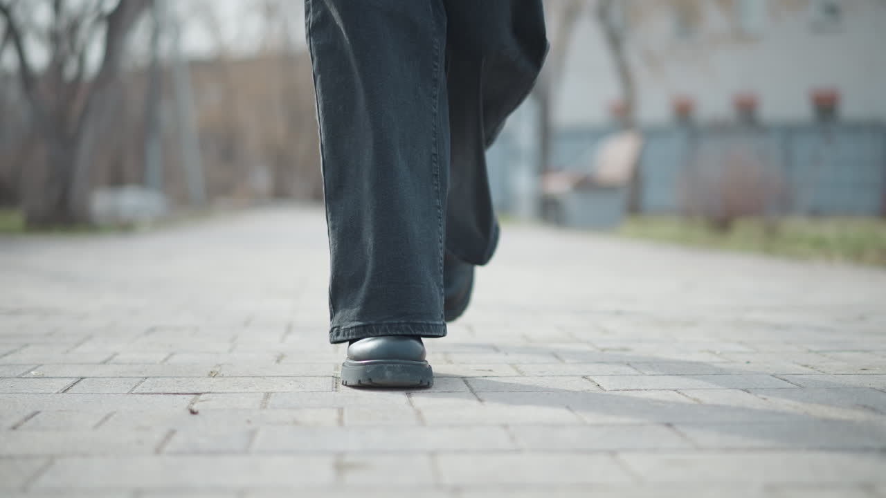 Close-up low-angle view of woman walking on paved park path in black pants and boots during cold winter day, capturing forward motion, footwear detail, and serene outdoor atmosphere with soft daylight