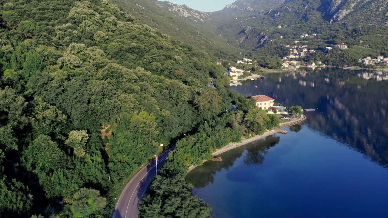 Camion che guida su una strada autostradale asfaltata che si snoda lungo la riva della baia di Cattaro, Montenegro, attraverso una piccola città costiera, con montagne e foreste circostanti che si riflettono nell'acqua, in una giornata di sole, ripresa aerea